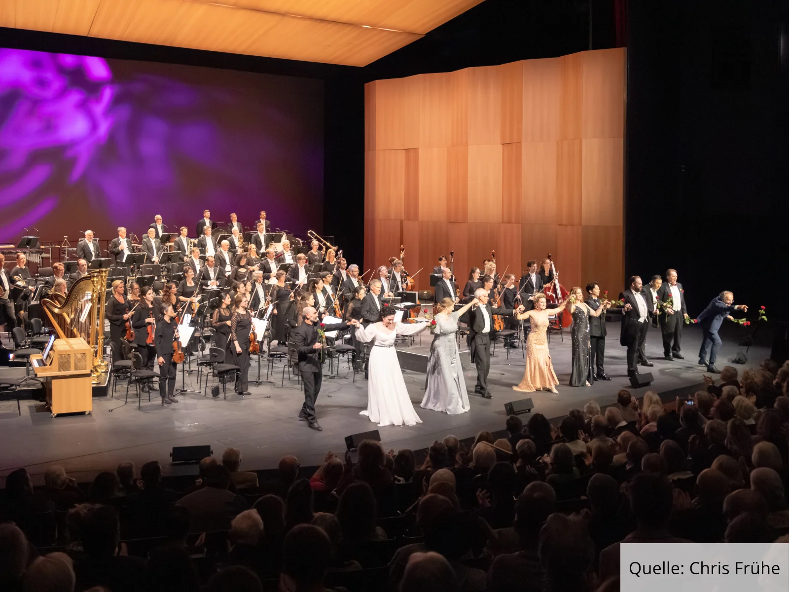 Ensemble und Orchester des Badischen Staatstheaters Karlsruhe beim Schlussapplaus auf der Bühne vor vollem Zuschauerraum